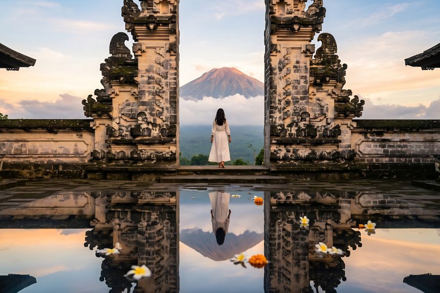 A lone traveller in a white dress stands between the ornate stone Gates of Heaven at Pura Lempuyang temple, with Mount Agung volcano perfectly reflected in the still pool below, Bali