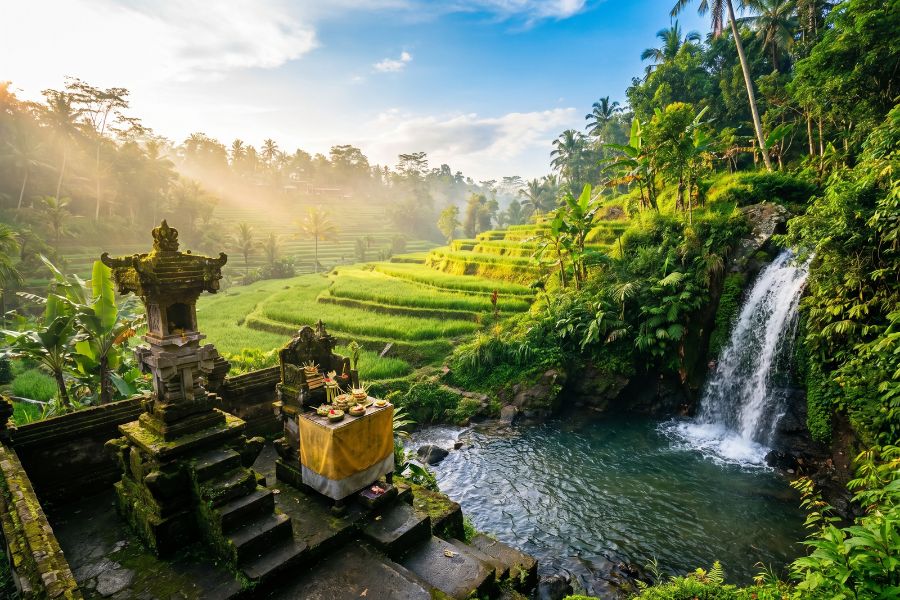 Emerald-green Tegalalang rice terraces in Ubud with a mossy water temple and jungle waterfall under a bright blue tropical sky.
