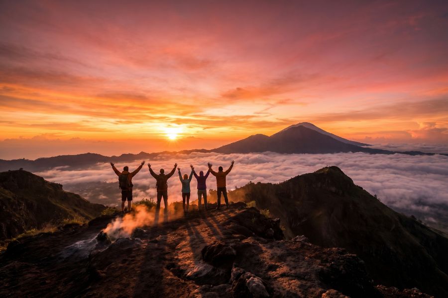Silhouetted hikers standing triumphantly on the volcanic crater rim of Mount Batur at sunrise, with a sea of white clouds below and Mount Agung on the horizon, Bali
