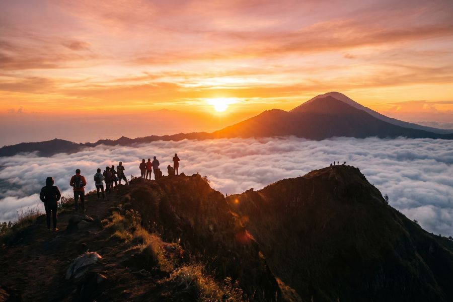Silhouetted hikers on the Mount Batur volcanic rim at sunrise, with Mount Agung rising above the clouds and a glowing orange sun over the caldera.