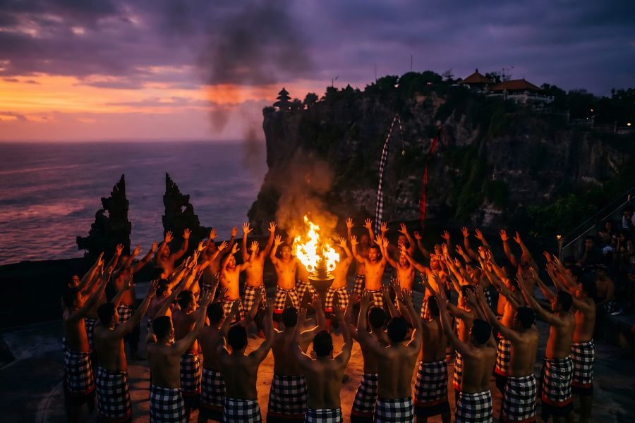 Dramatic low-light photograph of the Kecak fire dance at Uluwatu Temple, Bali, with fifty dancers chanting around a flaming torch and the clifftop sea temple silhouetted against a violet and orange sunset sky