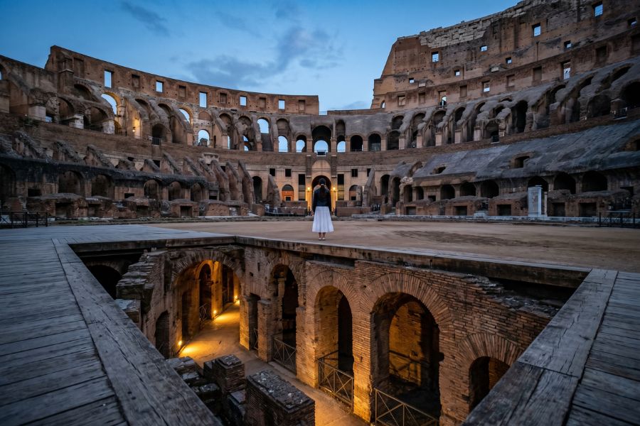 Immersive low-angle shot of a lone traveler on the arena floor looking down into the hypogeum of the Colosseum before sunrise.
